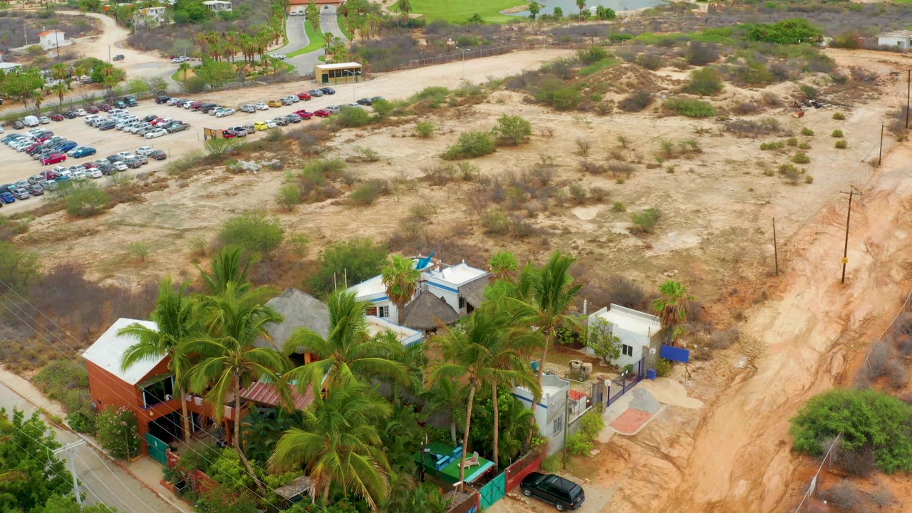 Drone view of dry terrain, parked cars, palm trees, rooftops, and a sandy road.