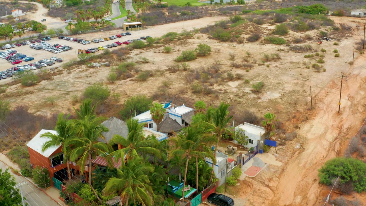 At 0:02, La Playita Los Cabos appears from above with dense palm trees and small structures in the lower third, dry scrubland across the center, a sandy road at right, a parking area at upper left, and no visible people.