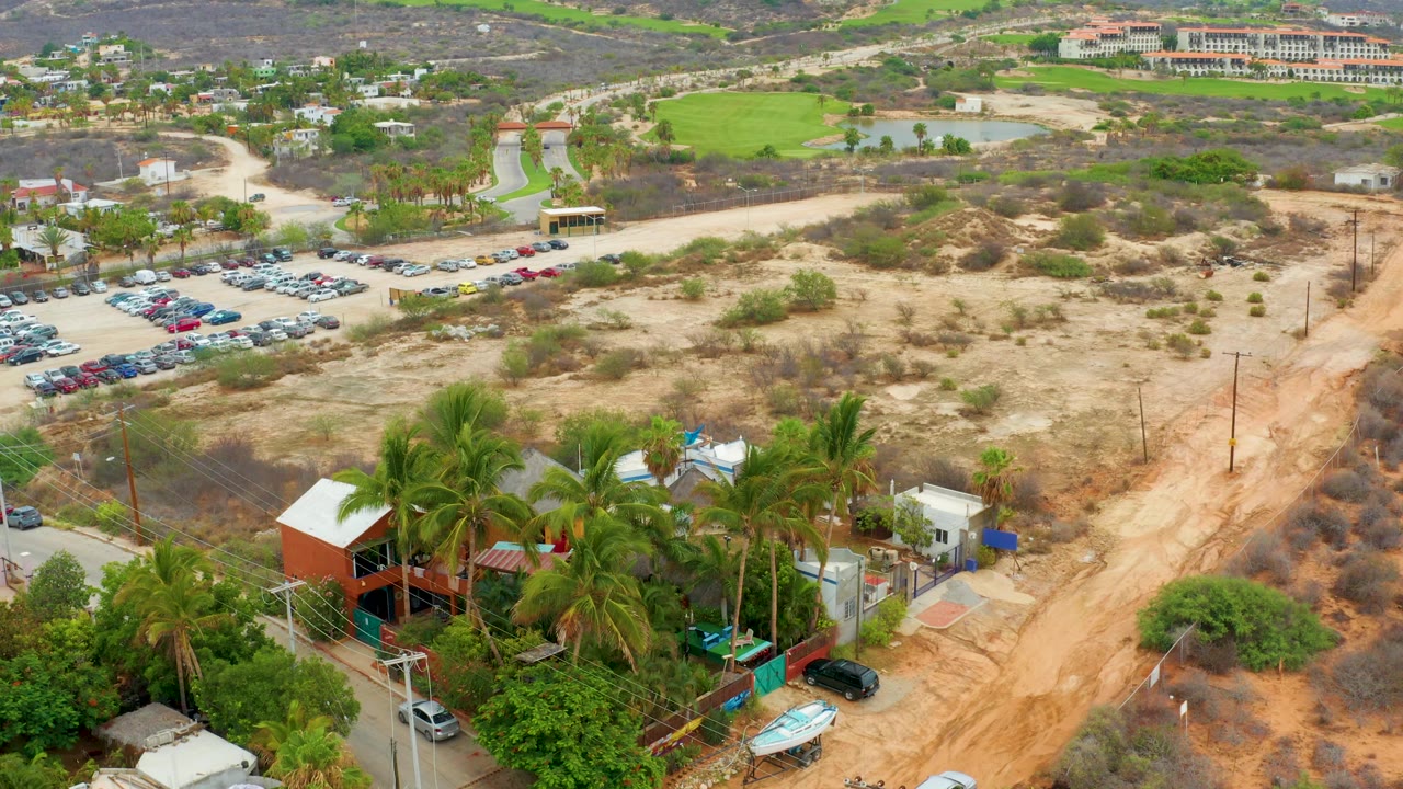 Aerial frame over La Playita Los Cabos showing a sandy dirt road, parked cars, palm trees, low buildings, scrub vegetation, and no visible people.