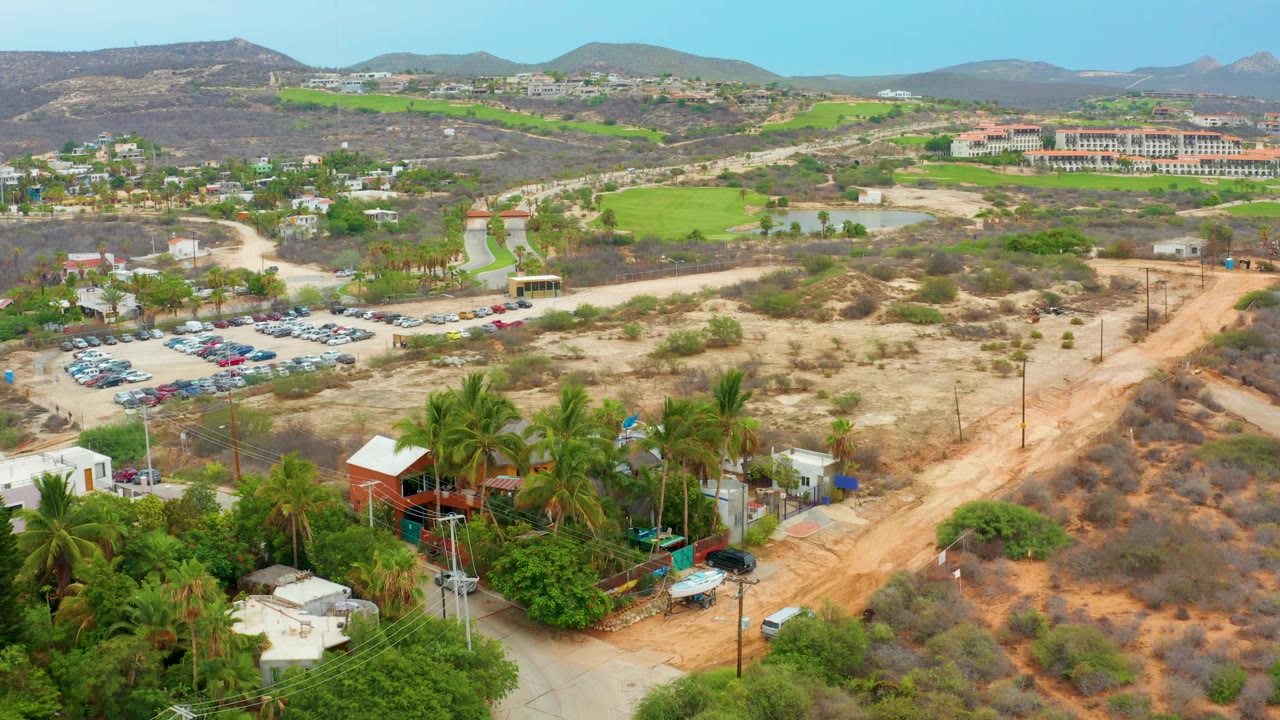 High drone view across palms, a dirt road, parked cars, green areas, and hills.