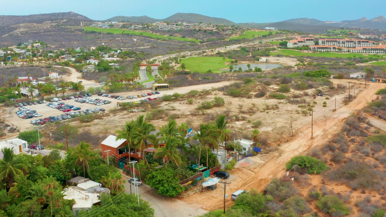 La Playita Los Cabos aerial frame with the dirt road running through dry scrub, parked vehicles to the left, palms and rooftops below, green patches beyond, and no visible people.