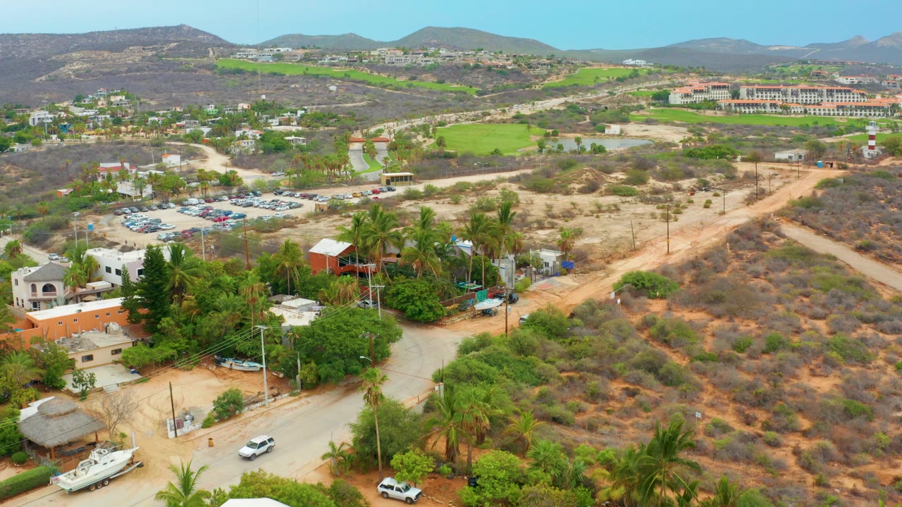 Aerial frame over La Playita Los Cabos showing dirt roads, palm trees, low buildings, a parking lot, dry scrub, green fields, and distant hills, with no visible people.