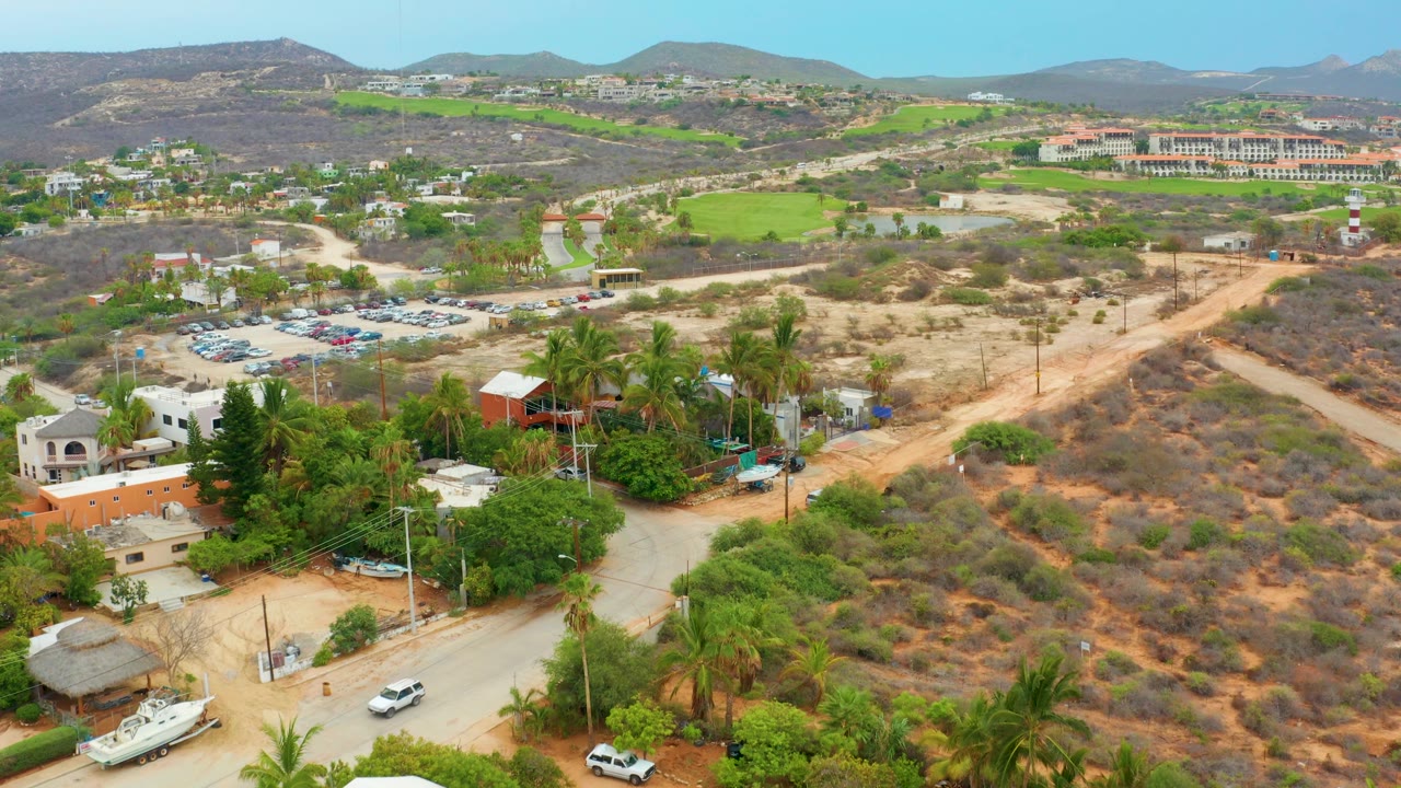 Aerial frame in La Playita Los Cabos with a road crossing the foreground near palms and buildings, a parking lot to the left, dry hillside scrub to the right, and no visible people.