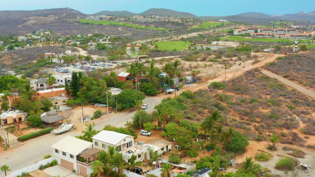Opening aerial frame over La Playita Los Cabos homes, palm trees, a pale road, dry scrubland, green fields, distant hills, and no visible people.