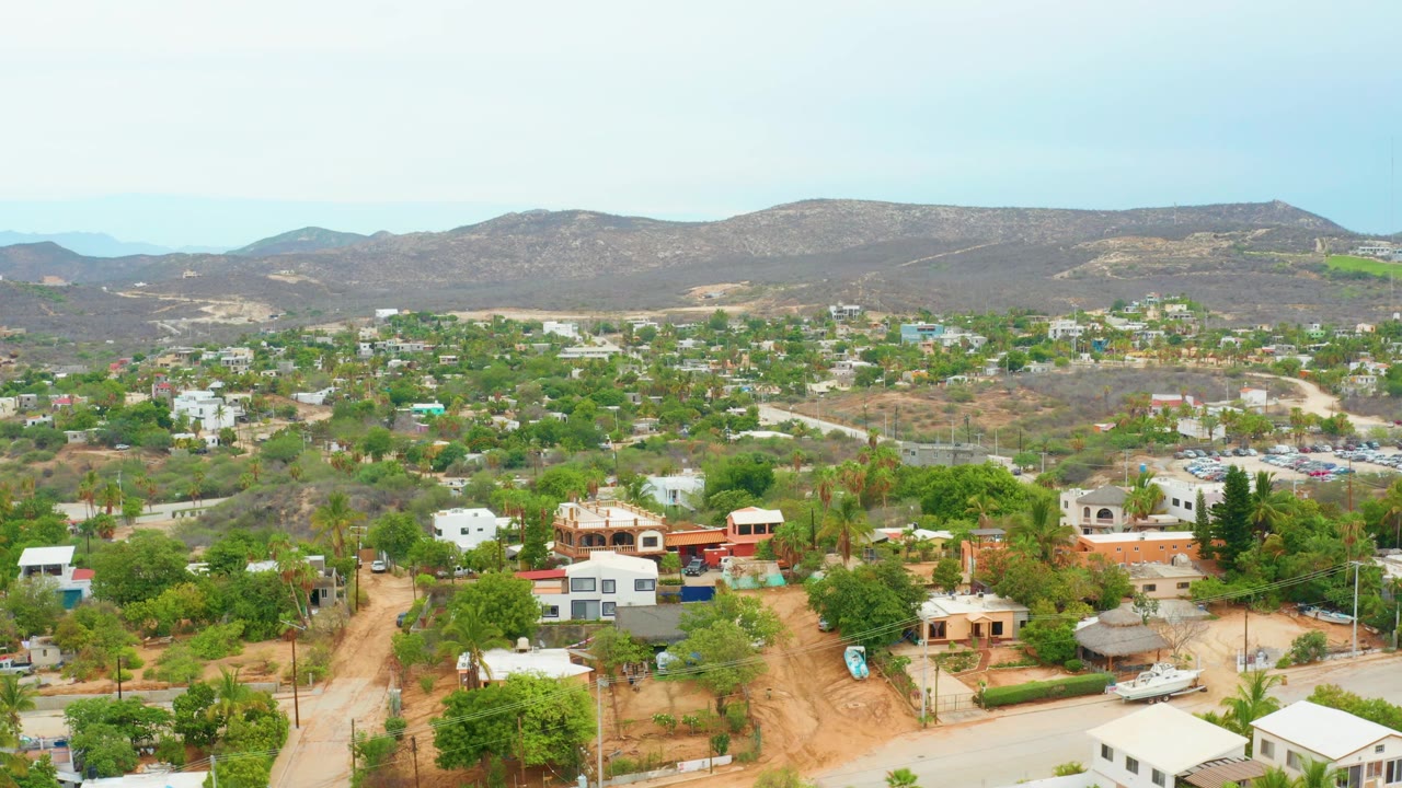 Aerial view over La Playita Los Cabos with scattered homes, green trees, dirt roads, dry hills, a pale overcast sky, and no visible people.
