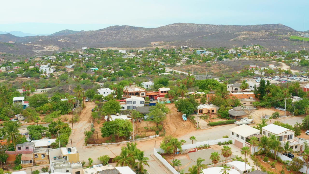 Opening aerial frame over La Playita Los Cabos showing residential houses, green trees, pale roads, dry mountain ridges, blue sky, and no visible people.