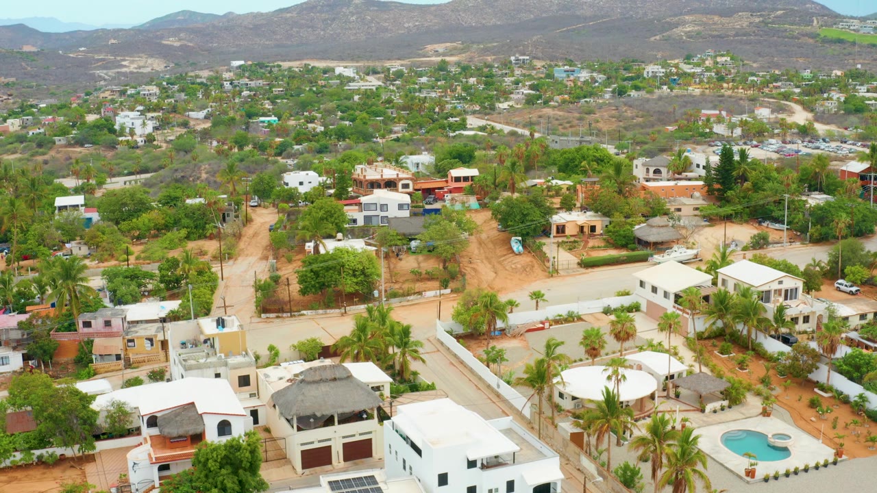 Aerial frame in La Playita Los Cabos with a turquoise pool near the lower center, white houses, palms, unpaved streets, patchy vegetation, arid slopes, and no visible people.