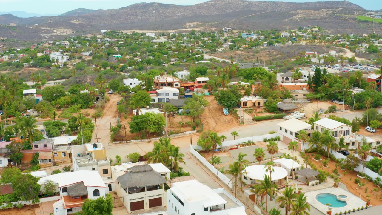 Aerial frame over La Playita Los Cabos showing the neighborhood spread across green and tan terrain, with flat-roof homes, palms, winding roads, mountains, and no visible people.