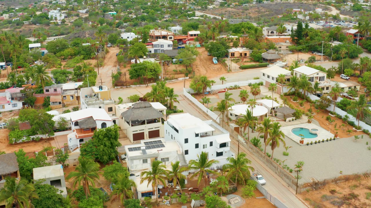 Aerial frame of La Playita Los Cabos showing white houses, palm trees, a road grid, dry reddish ground, a small blue pool, distant hills, and no visible people.