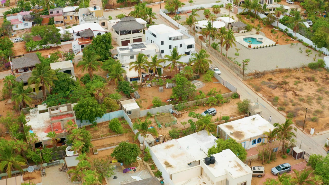 High aerial frame over La Playita Los Cabos showing white homes, palm trees, sandy yards, a street, and no visible people at the start of the clip.