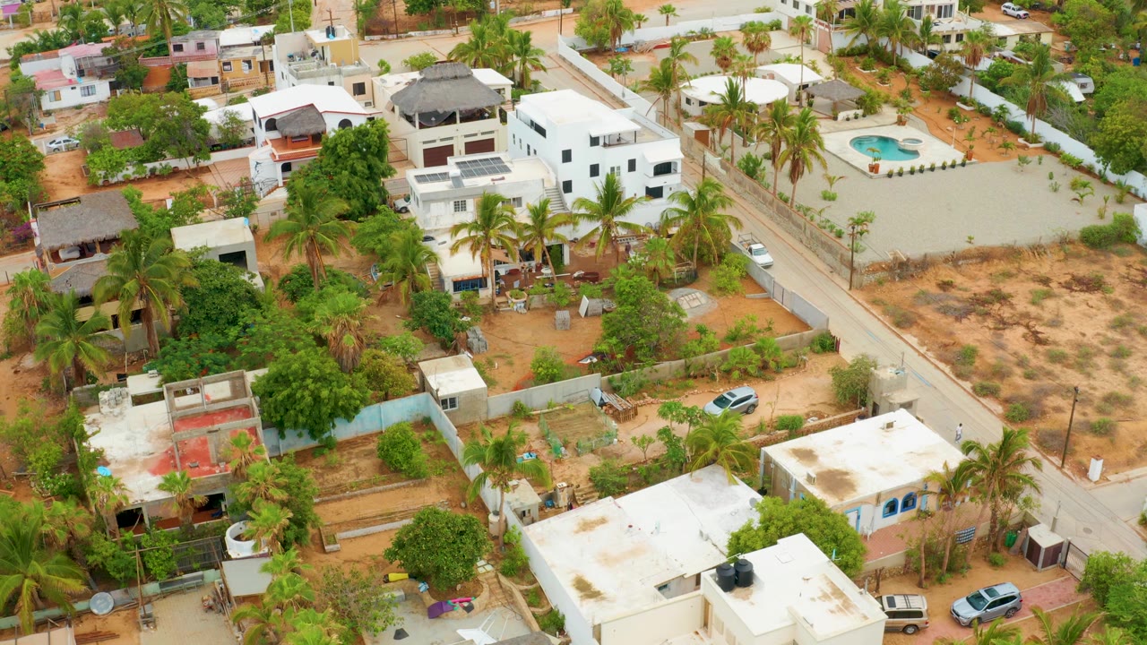 High oblique frame over La Playita Los Cabos showing the road running between white houses, palm-lined yards, a dry open lot, parked vehicles, and no visible people.