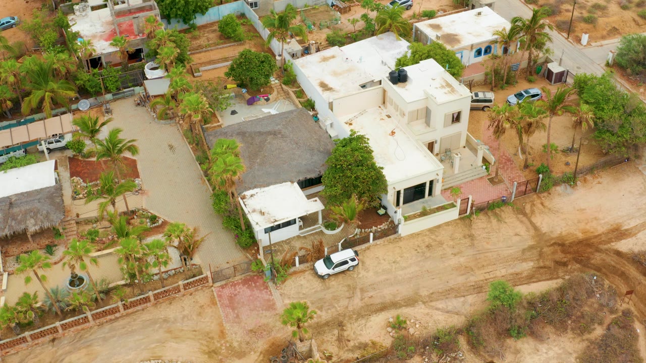 Aerial view over La Playita Los Cabos showing white flat-roof homes, palm trees, walled yards, a dirt road, and dry brown ground with no visible people.