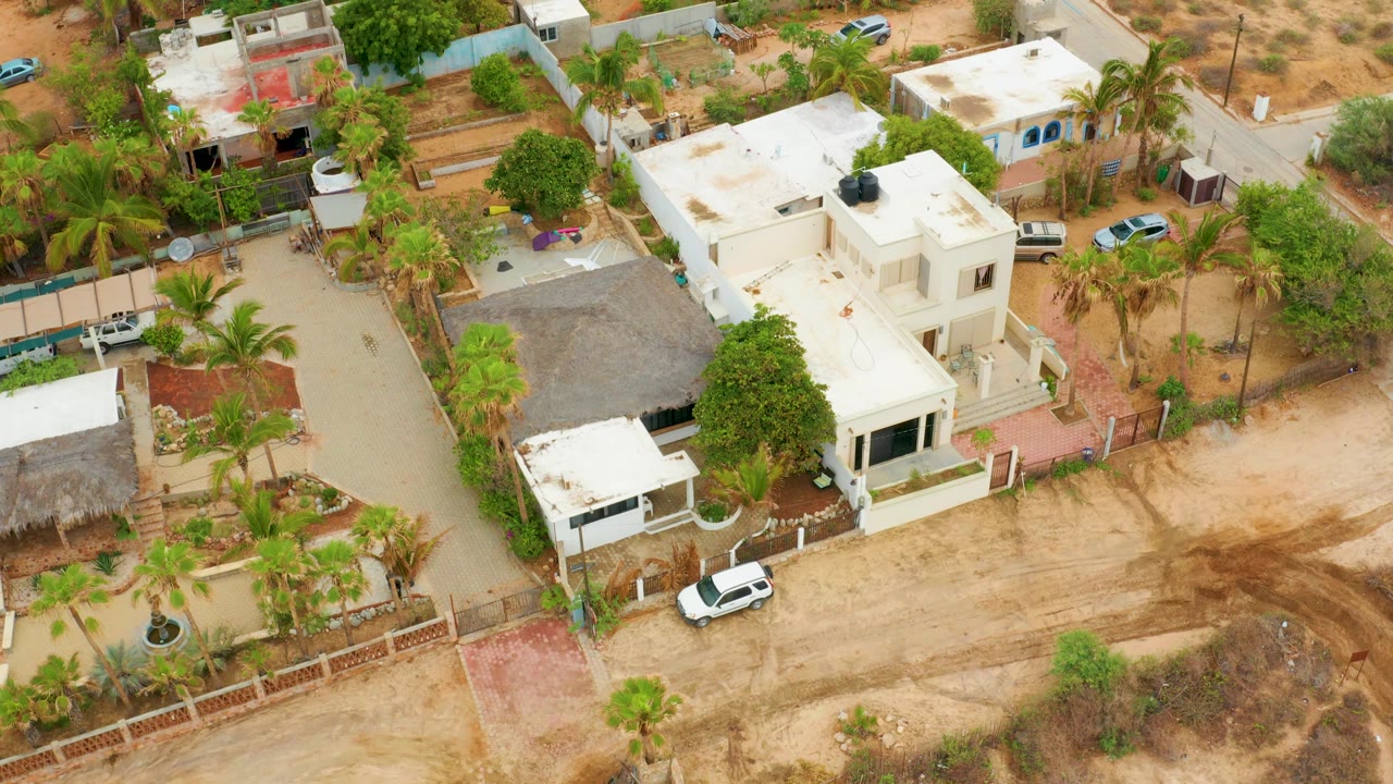 Oblique aerial frame over La Playita Los Cabos showing the residential lane, white homes, palms, a thatched-looking roof section, parked vehicles, and no visible people.