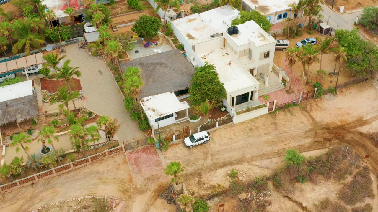 Aerial frame over La Playita Los Cabos showing white flat-roof homes, palms, dirt lots, fenced yards, and a parked white car with a small person near the thatched structure.