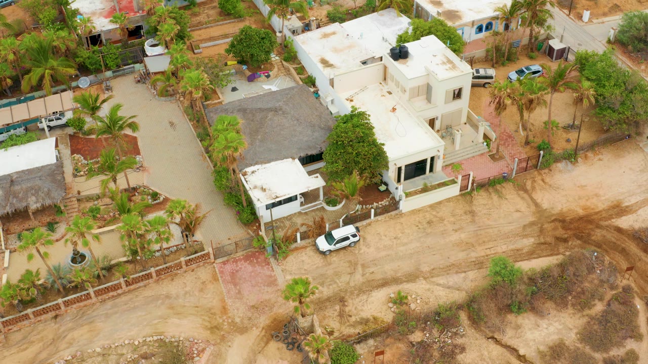 High oblique frame over La Playita Los Cabos showing the white house cluster, palm-lined yards, unpaved roads, dry vacant ground, and the same parked white car near the lower edge.