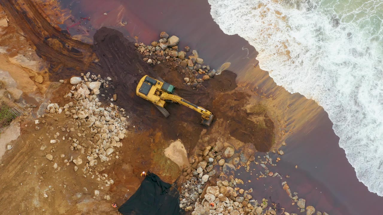 Overhead frame of La Playita Los Cabos with the yellow excavator angled across the center, rock piles on both sides, brown water around the machine, surf entering from the upper right, and no visible people.