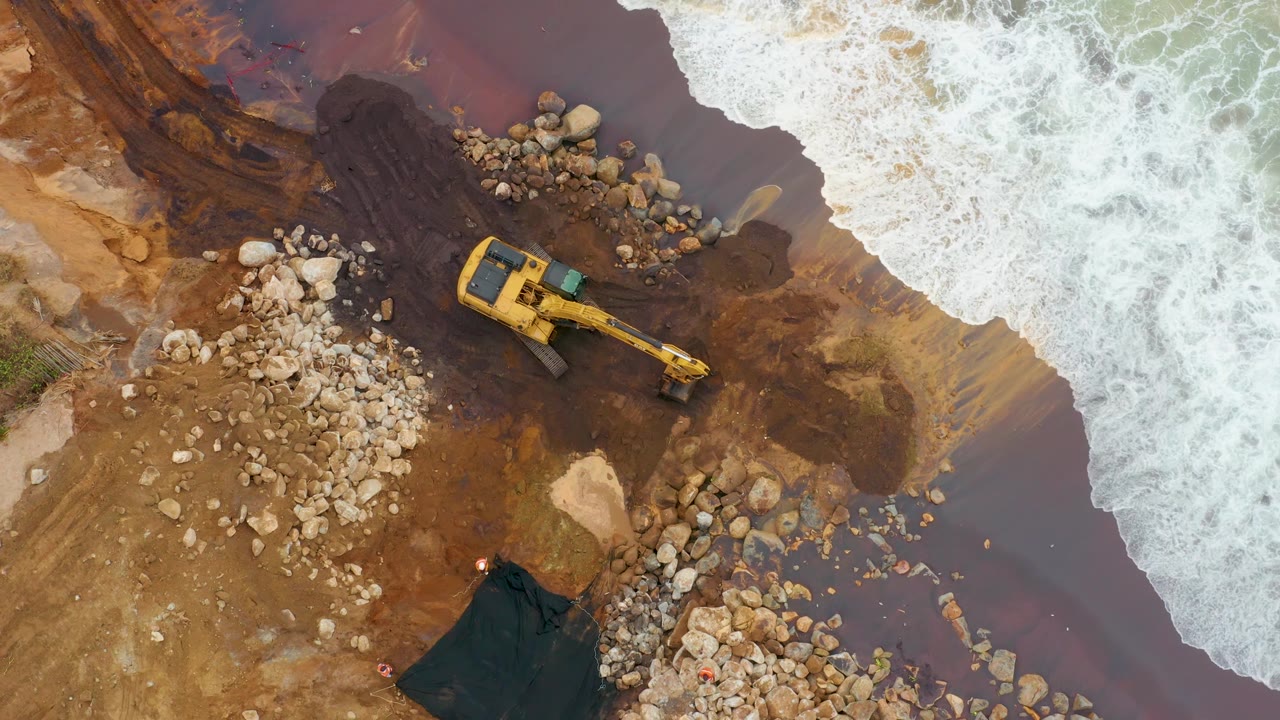 Overhead frame of La Playita Los Cabos where the excavator arm extends toward the lower right rocks, foamy water fills the upper right, brown runoff colors the shore, and no visible people appear.