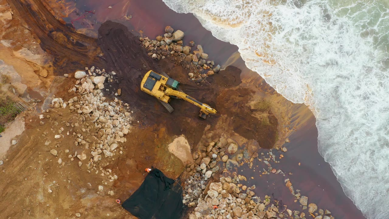 Excavator on rocky shoreline as a larger wave reaches the work area.