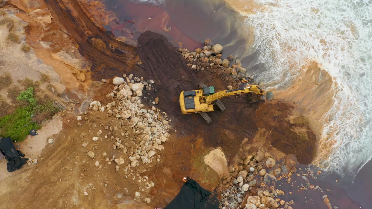 Aerial view in La Playita Los Cabos with the excavator arm reaching toward the shoreline, surrounded by boulders, dark brown water, tan sand, and foamy waves, with no visible people.
