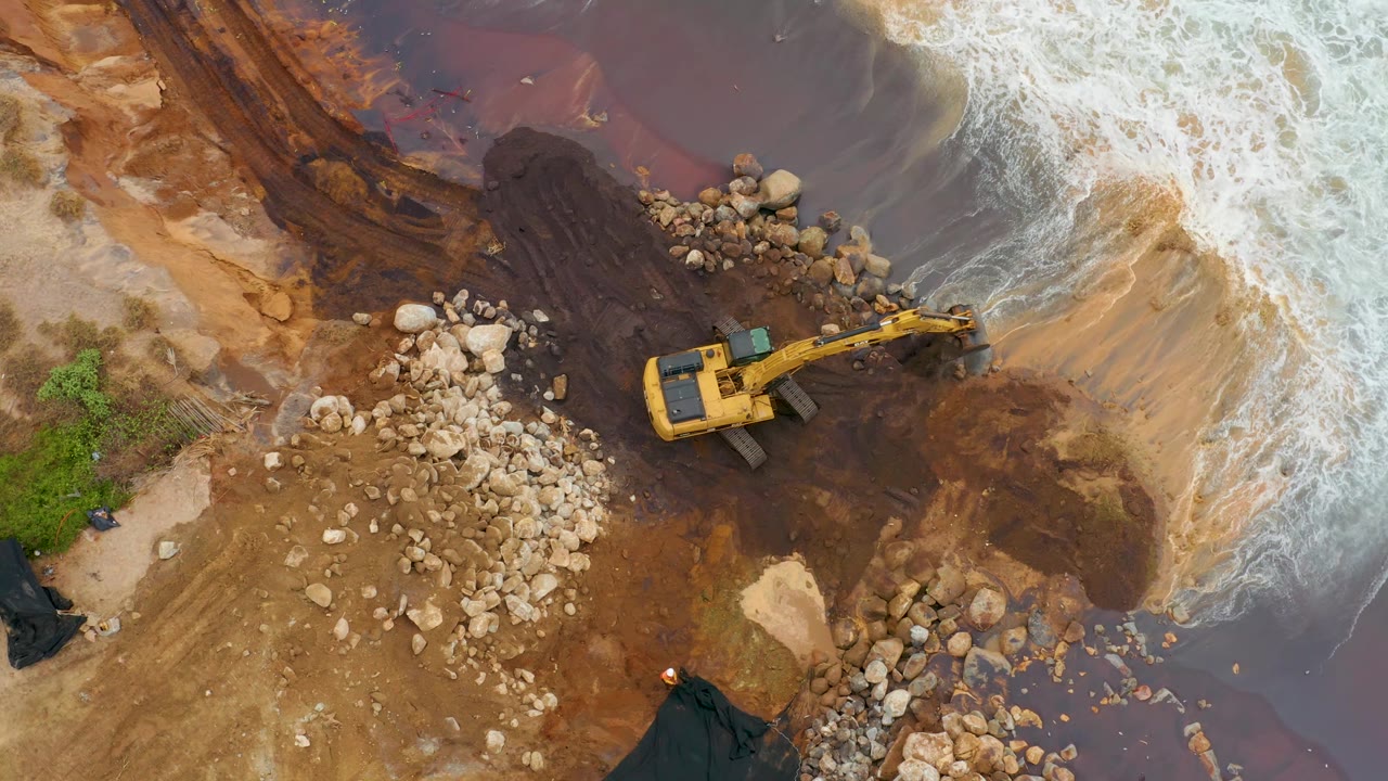 La Playita Los Cabos overhead frame showing the excavator shifted near the right side of the rock field, with dark runoff-like water and white surf patterns around the shore, with no visible people.