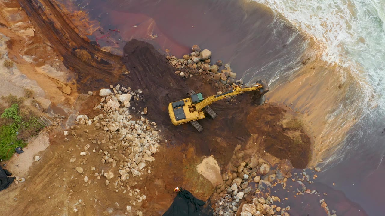 Excavator on brown rocky beach with surf entering from the upper right.