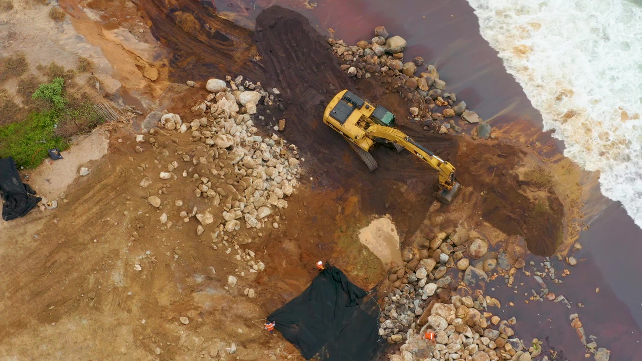 Overhead drone frame in La Playita Los Cabos showing a yellow excavator near pale rock piles, brown muddy ground, white surf, black tarps, and no visible people.