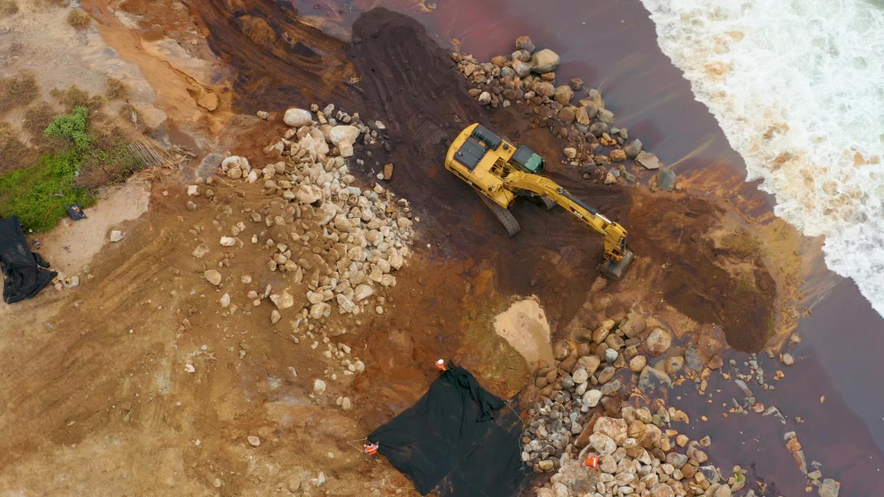 Aerial frame in La Playita Los Cabos with the excavator arm extended toward rocks beside dark muddy water, white surf at the right edge, and no visible people.