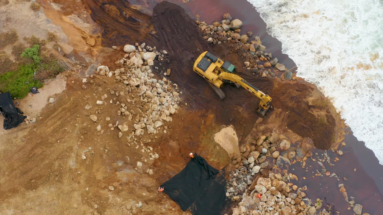 High overhead view in La Playita Los Cabos showing the excavator slightly higher in frame beside a dark muddy flow, scattered rocks, breaking white waves, and no visible people.