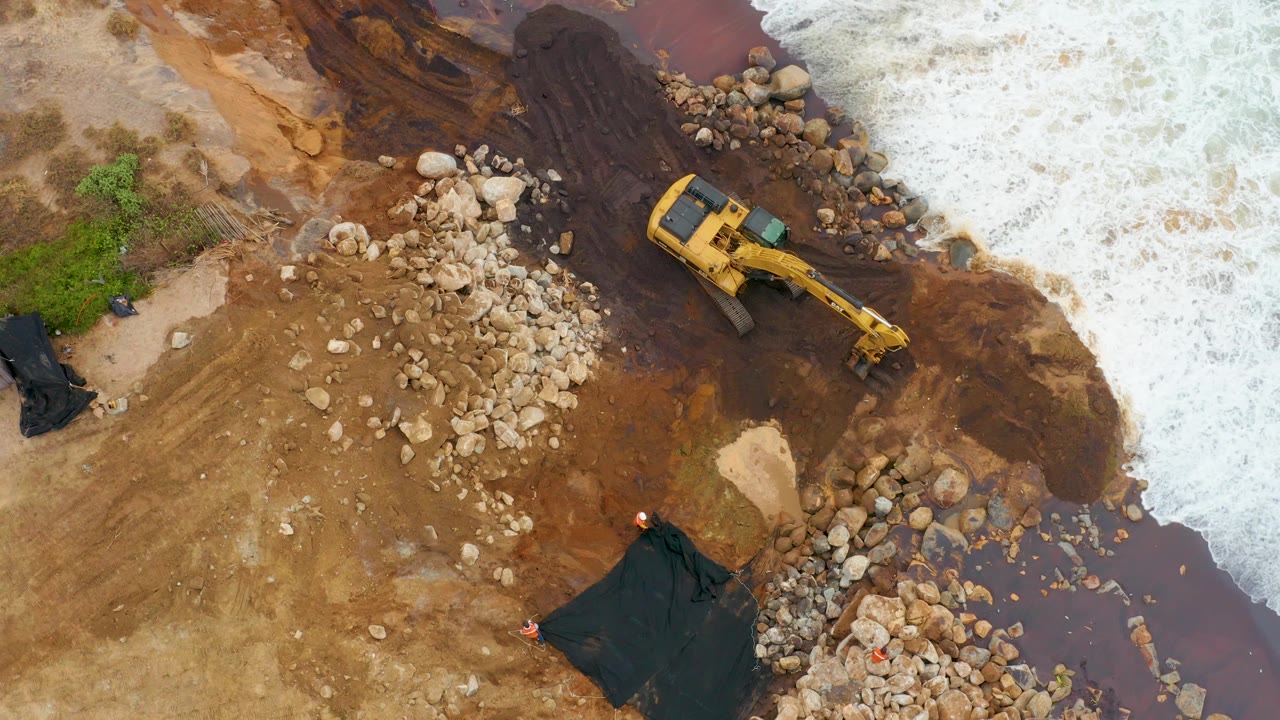 Aerial frame in La Playita Los Cabos where the excavator is centered beside rock piles and muddy brown water while white surf pushes in from the right, with no visible people.