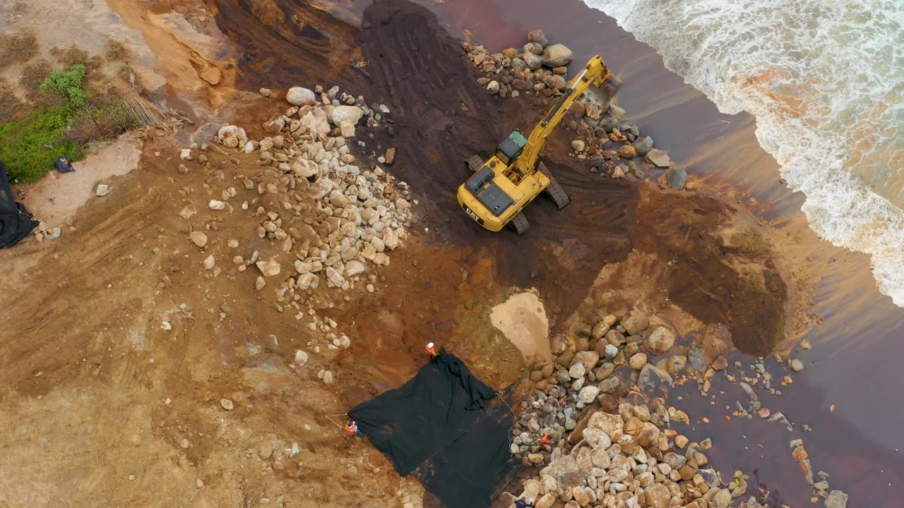 Overhead frame from La Playita Los Cabos showing a yellow excavator on wet brown mud between pale rock piles and white surf, with no visible people.