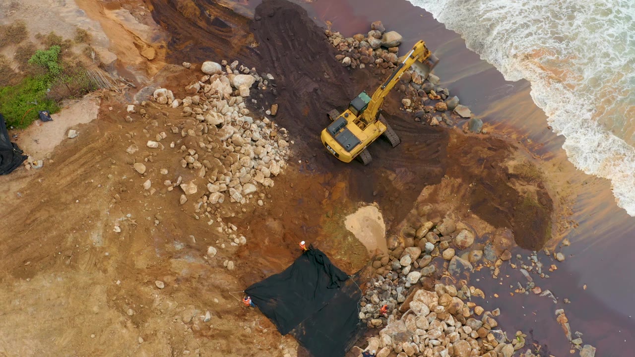 At 0:01, the excavator remains near the right side of the muddy La Playita Los Cabos shore while ocean foam curls along the upper-right edge and no people are visible.