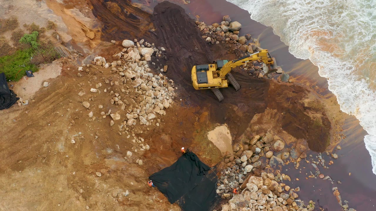 At 0:02, the excavator arm extends toward a cluster of shoreline rocks while surf presses in from the right on the muddy La Playita Los Cabos coast, with no visible people.
