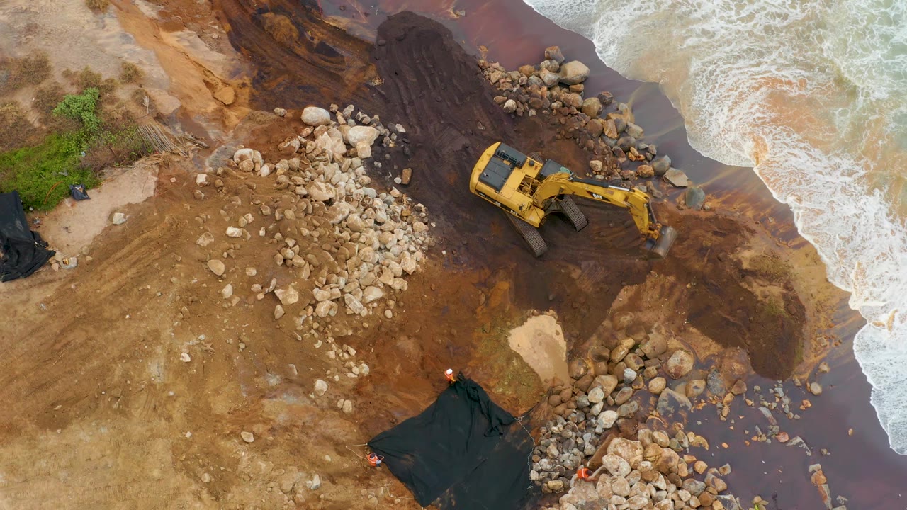 At 0:03, the yellow excavator sits beside the central pale rock pile on wet brown mud in La Playita Los Cabos, with foamy water entering from the right and no visible people.