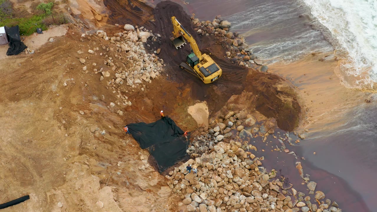 Aerial frame in La Playita Los Cabos, Mexico, with the excavator slightly farther along the shoreline, rock piles below it, foamy surf at right, and a black tarp near the lower center, with no visible people.