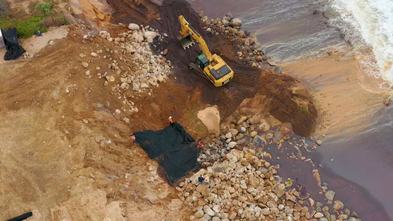 High overhead frame in La Playita Los Cabos, Mexico, showing the excavator near the upper right, dark mud leading to the water, rock piles across the shore, and surf pushing in from the right, with no visible people.