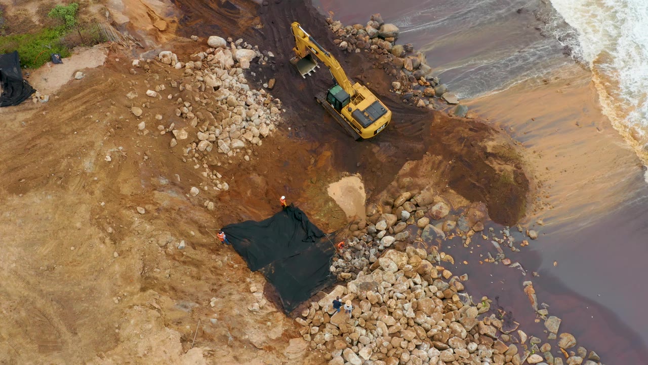 Aerial view in La Playita Los Cabos, Mexico, showing the excavator near the water's edge, a black tarp lower left, large rocks across the lower shore, and pale surf at the right edge, with no visible people.