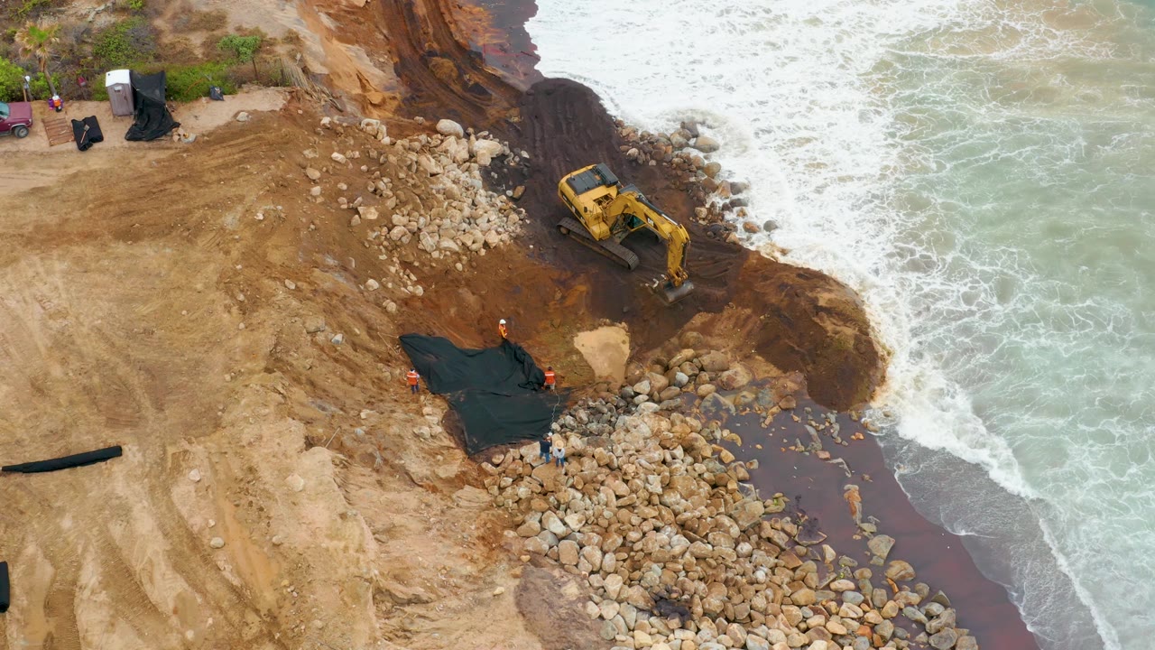 Aerial view in La Playita Los Cabos, Mexico, with the excavator angled near a pile of boulders, dark fabric below it, and white waves pushing against the shoreline, with no visible people.