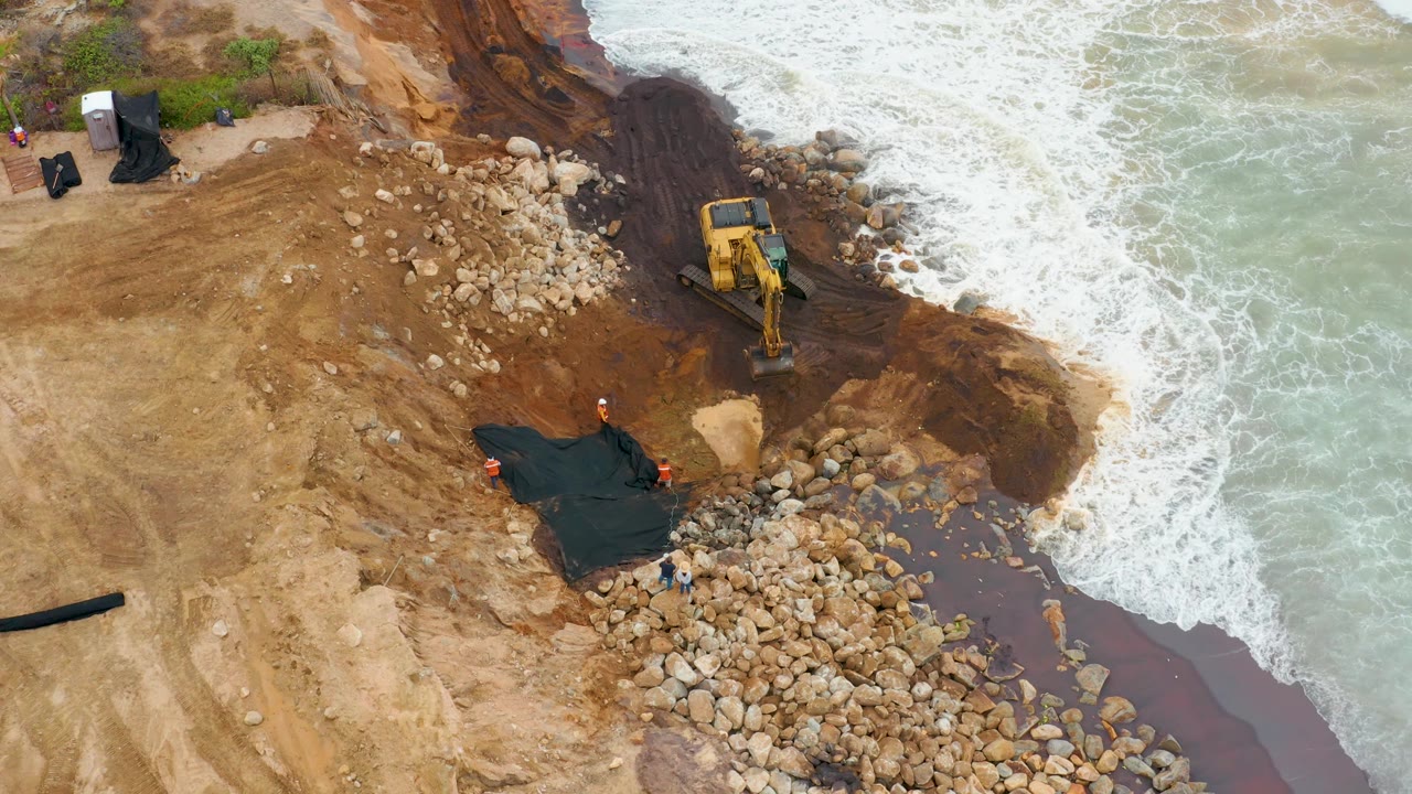 Excavator arm over rocks as waves wash along the shoreline construction site.