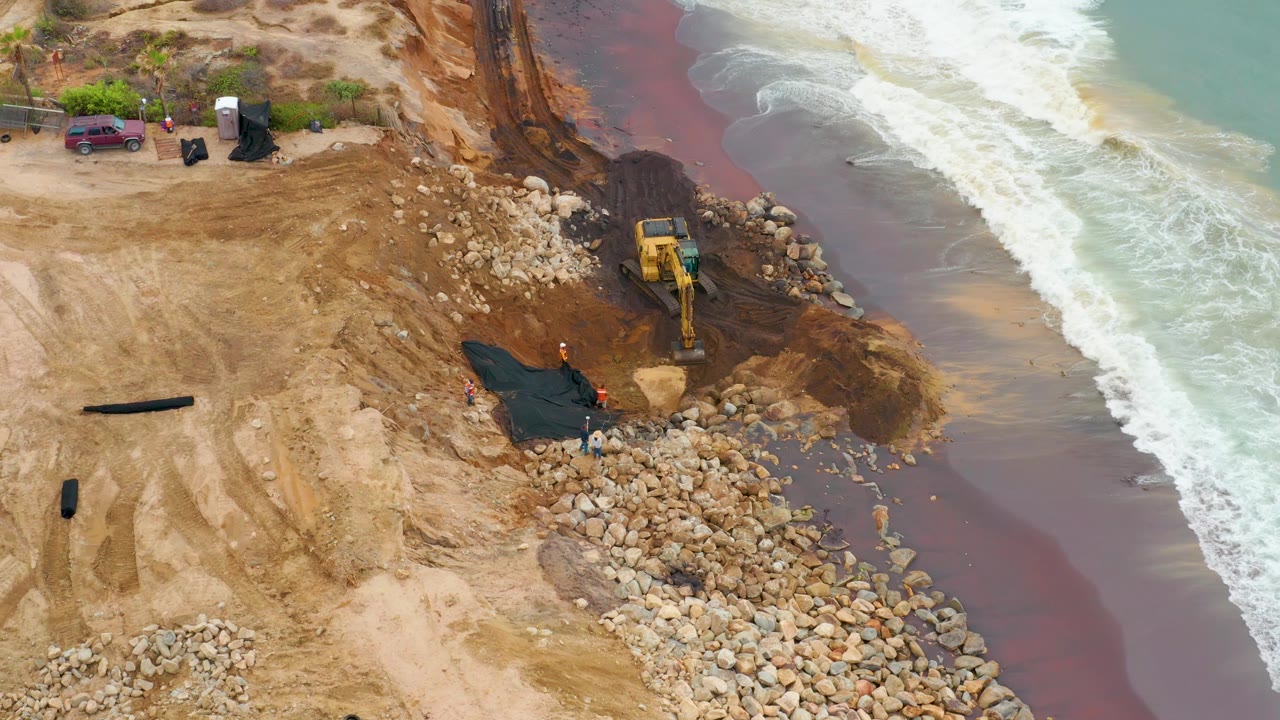 Aerial view in La Playita Los Cabos with the excavator bucket near dark soil, large rocks spread along the beach, white surf curling at the right edge, and no visible people.