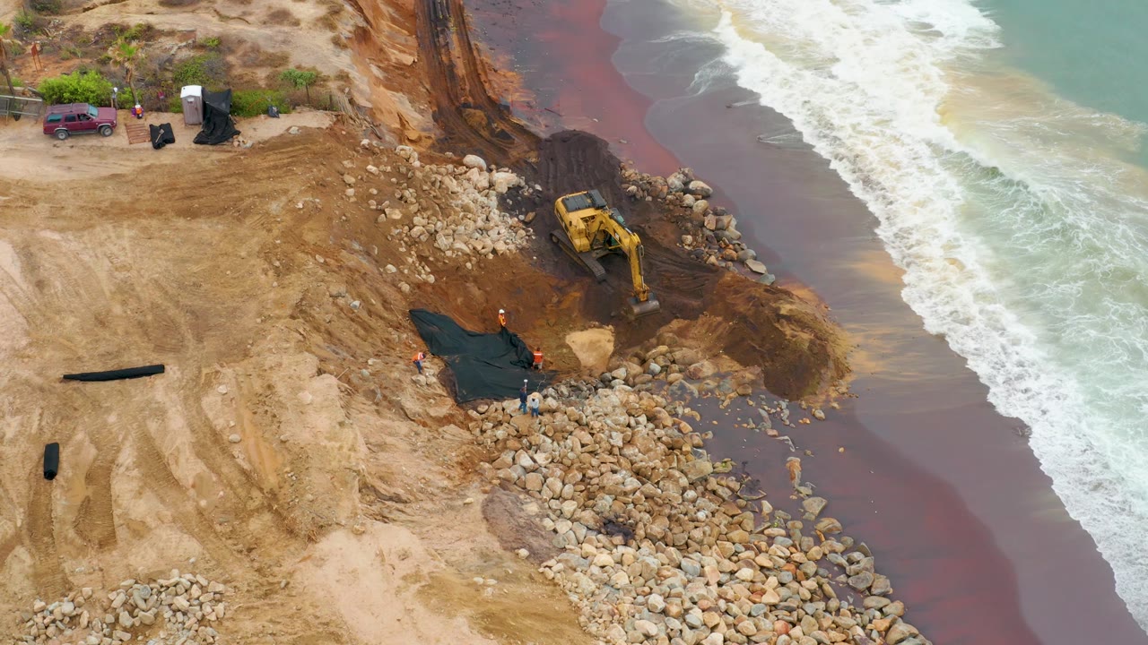 La Playita Los Cabos aerial frame showing the excavator beside a cut in the shoreline, pale green water and white foam on the right, tan graded sand on the left, and no visible people.