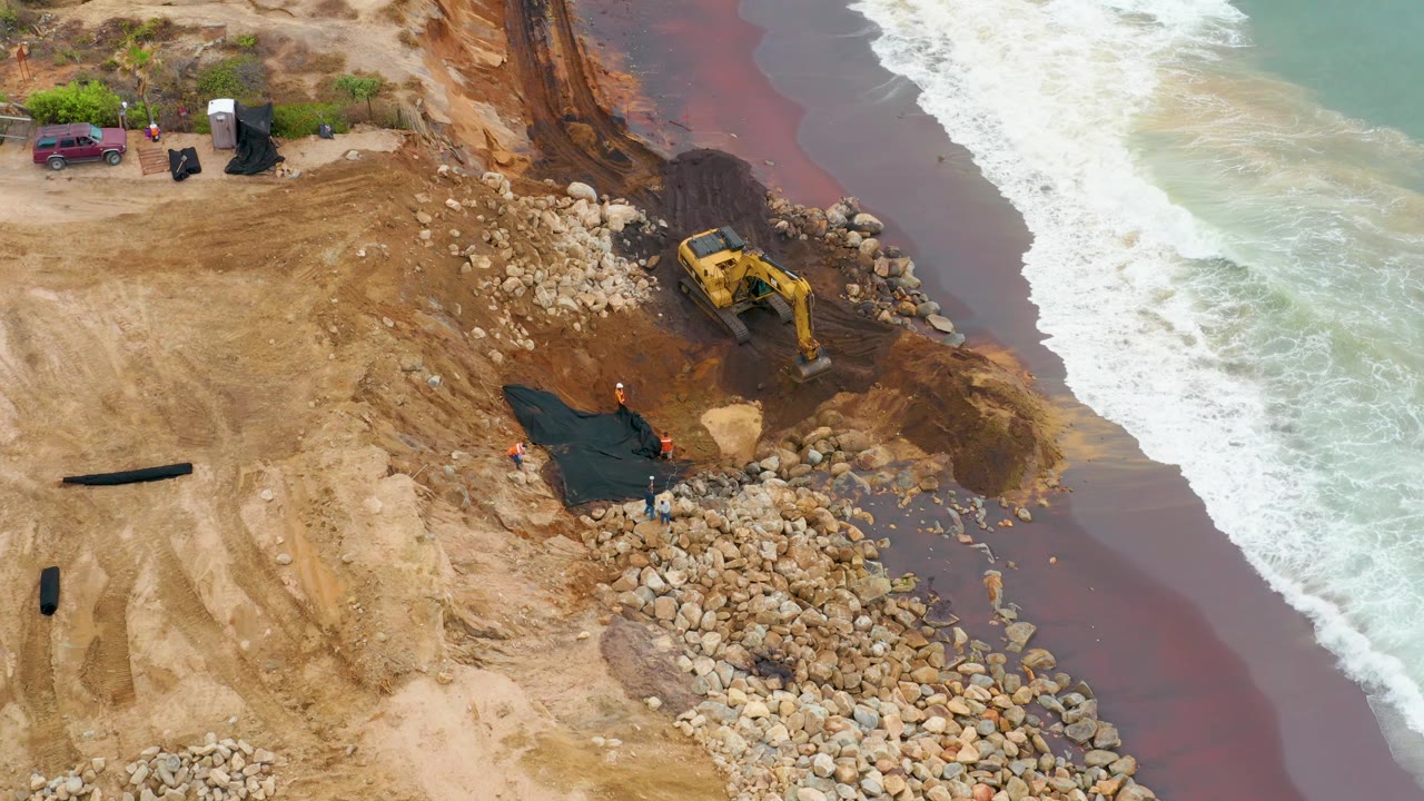Aerial frame in La Playita Los Cabos with the excavator angled toward the surf, rocks covering the lower shoreline, a dark tarp-like sheet on the sand, and no visible people.
