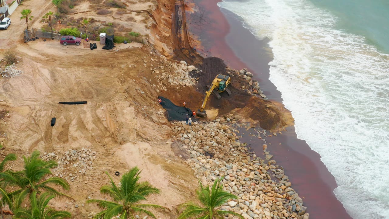 Aerial frame from La Playita Los Cabos, Mexico, showing a yellow excavator beside a rock berm where white surf meets tan sand, with palms at the lower edge and no recognizable people visible.