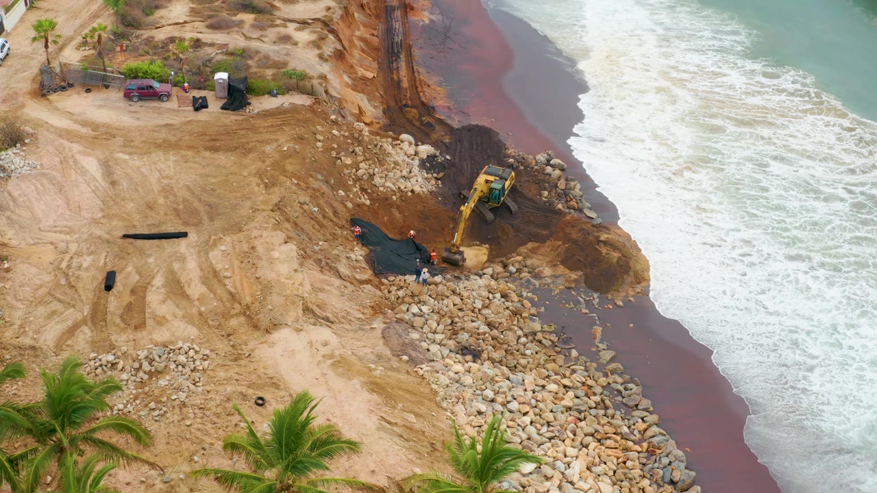 Aerial frame from La Playita Los Cabos, Mexico, showing foamy surf pushing farther up the right edge while the excavator remains beside the rock barrier and no recognizable people are visible.