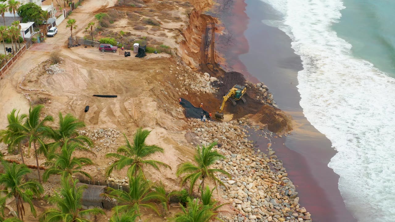 Aerial view in La Playita Los Cabos shows the excavator near the waterline, a broad sandy work pad, palm crowns in the lower frame, and no visible people.