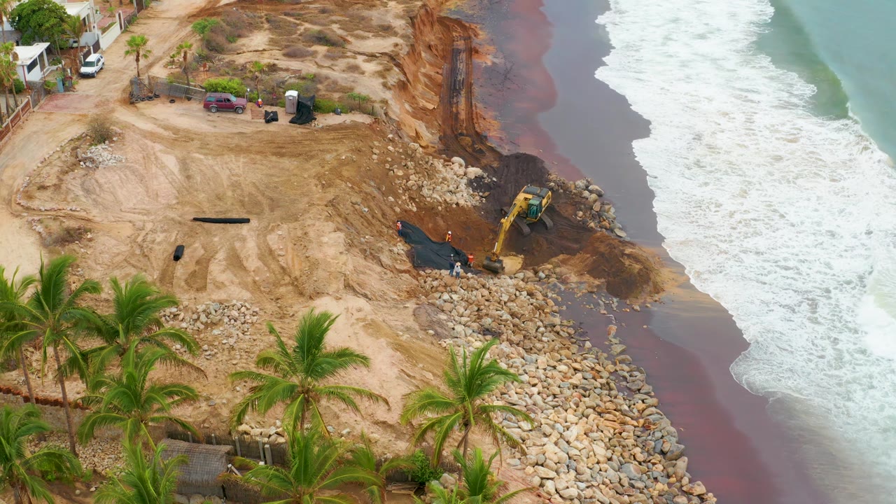 Aerial shoreline construction site with excavator and foamy waves.