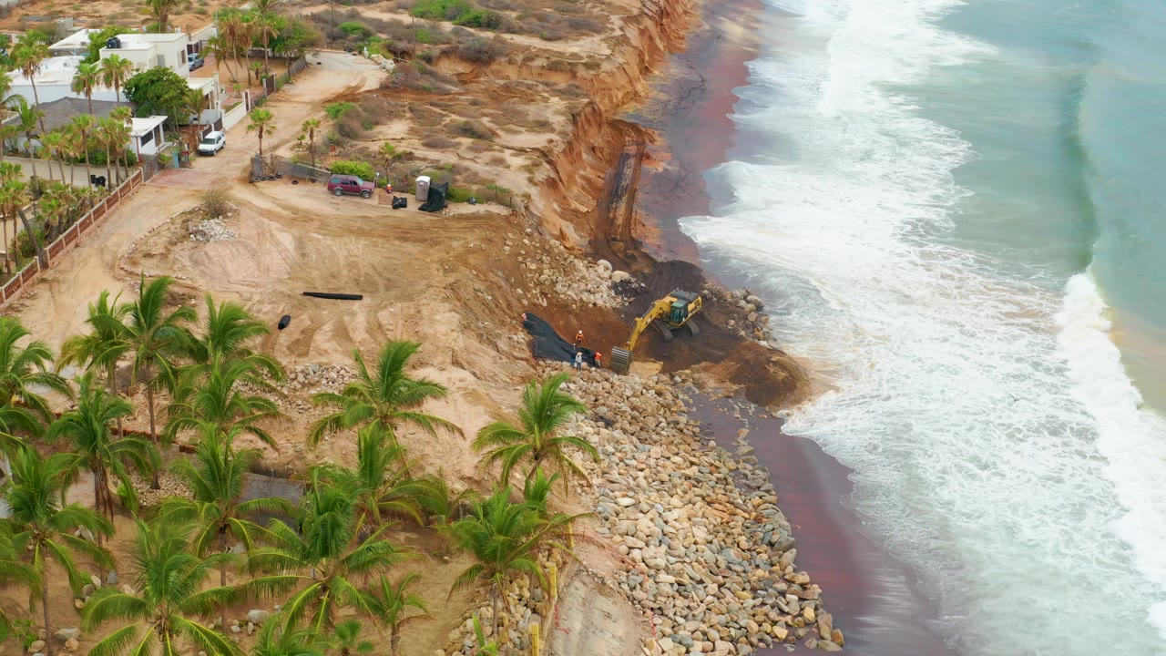 Aerial view in La Playita Los Cabos, Mexico, showing a yellow excavator beside an eroded cliff, rock protection, palm trees, coastal houses, and breaking surf, with no visible people.