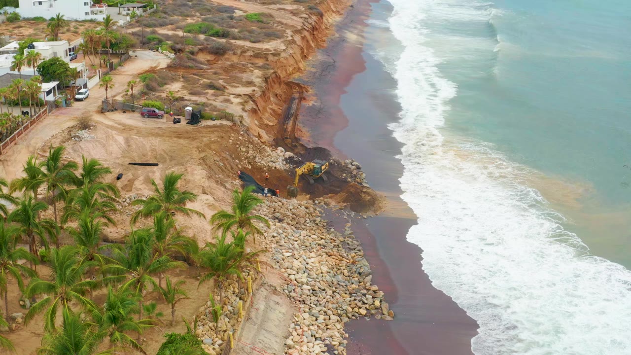 Aerial frame from La Playita Los Cabos showing palms and sandy ground beside an eroded cliff, a yellow machine near rocks, breaking surf at right, and no visible people.