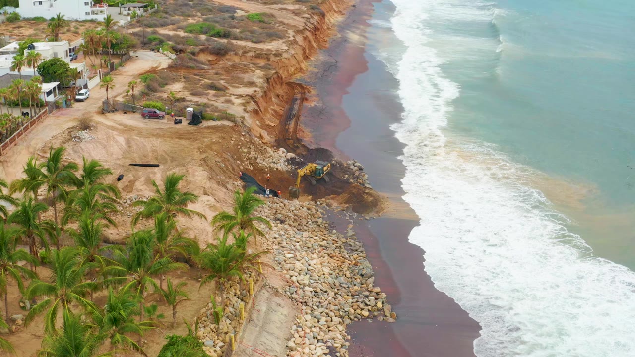 Drone-like aerial view in La Playita Los Cabos with the surf line slightly farther up the beach, the eroded cliff face exposed, palms at lower left, and no visible people.