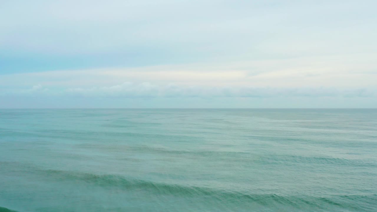 Turquoise-blue water fills most of the frame at La Playita Los Cabos, Mexico, with faint wave bands below a light gray cloud layer and no visible people.