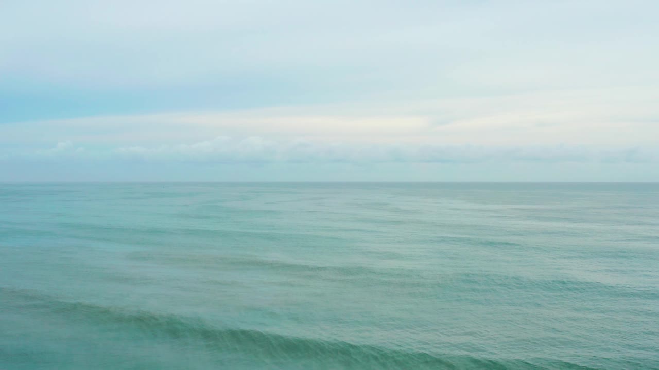 A wide elevated view shows open blue-green water at La Playita Los Cabos, Mexico, with a level horizon, muted cloud cover, and no visible people.
